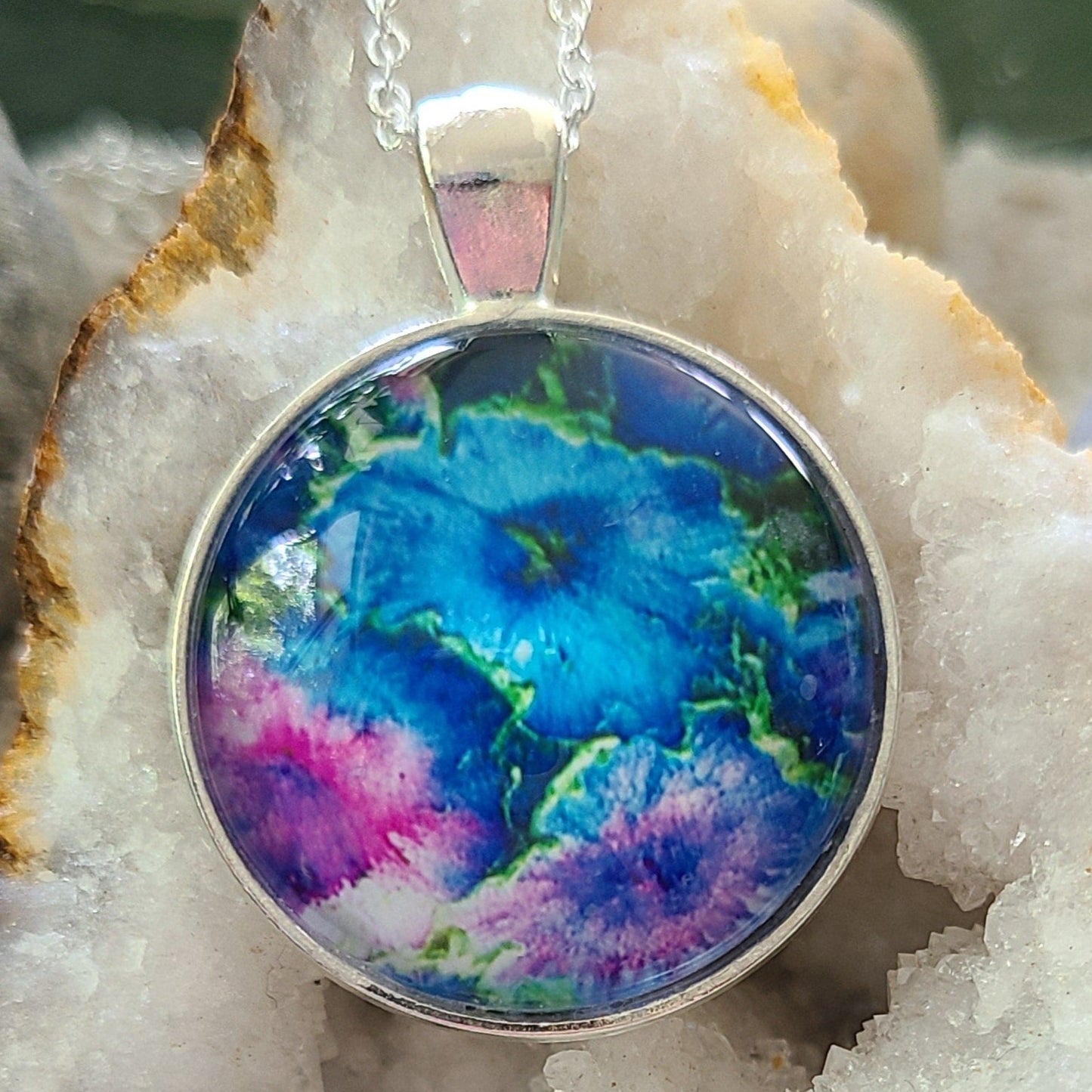 Alisa Marie original artwork pendant, My Favorite Things featuring ocean blue, green, and purple tones reminiscent of flowers, set in silver frame with silver chain, displayed on white crystal geode