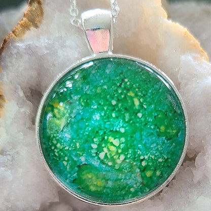 Alisa Marie original artwork. Emerald Dreams pendant featuring shimmering emerald green with yellow highlights, set in silver frame with silver chain, displayed on white crystal geode.