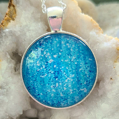 Alisa Marie original artwork, Healing Rain pendant featuring shimmering sky blue tones, set in silver frame with silver chain, displayed on white crystal geode.