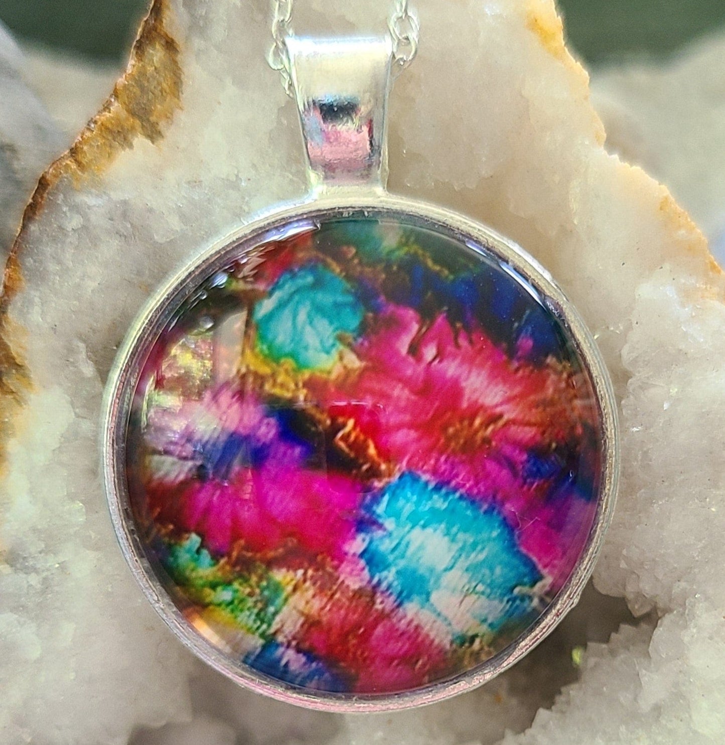 Alisa Marie original artwork,The Grateful Flower pendant featuring vibrant pink, turquoise, and rainbow colors, set in silver frame with silver chain, displayed on white crystal geode.