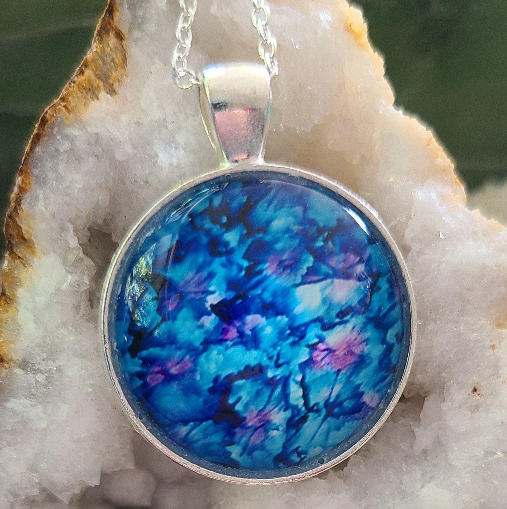 Alisa Marie original artwork, Dancing with Butterflies, pendant featuring vibrant ocean blues with purple accents, set in silver frame with silver chain, displayed on white crystal geode.