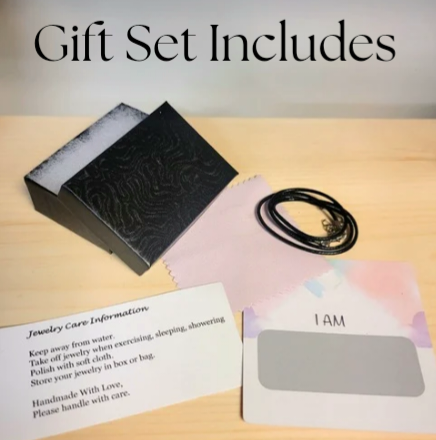 Alisa Marie Gift Set Includes: Black embossed jewelry box, black cord necklace, jewelry care information card with handling instructions, and 'I AM' affirmation card on wooden surface.
