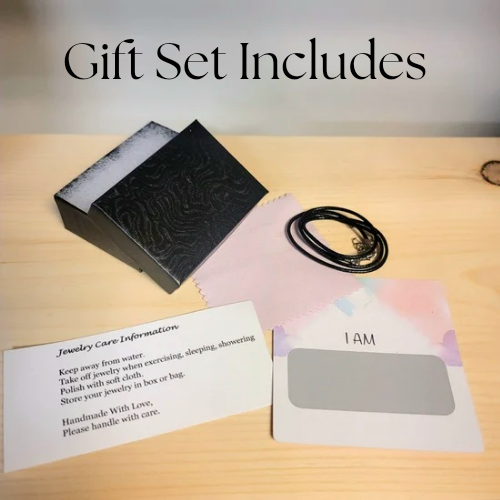 Alisa Marie Gift Set Includes: Black embossed jewelry box, black cord necklace, jewelry care information card with handling instructions, and 'I AM' affirmation card on wooden surface.
